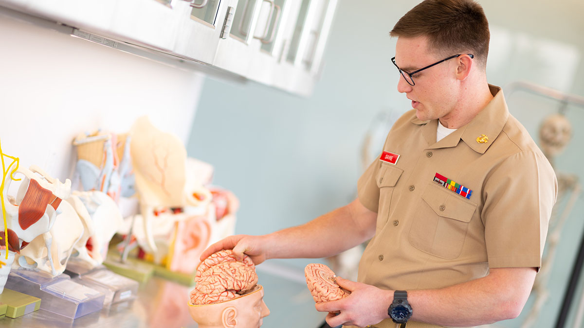 Young man wearing glasses and military blouse with ribbons and slacks stands in front of cupboard of anatomy models.