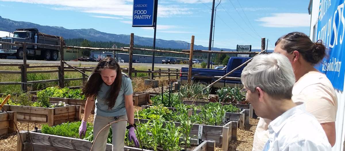 A woman with work gloves stands in a garden with signs reading "Cascade Food Pantry."