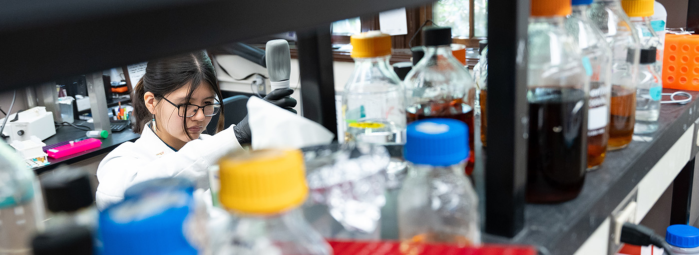 Woman standing behind a shelf full of colorful bottles in lab measures liquid with a pipette.