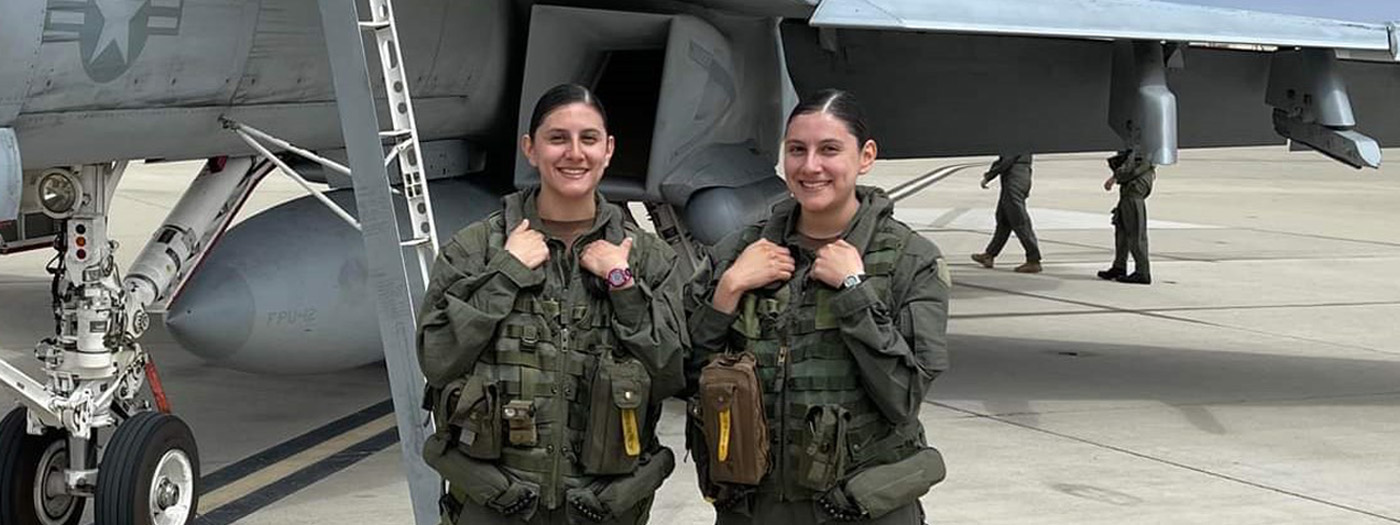 Two women in flight suits stand on tarmac next to a plane