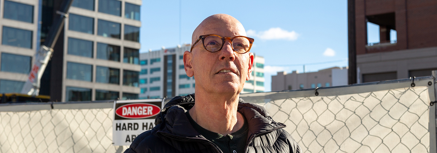 Jaap Vos poses in front of a construction zone fence with a crane and tall buildings in downtown Boise.