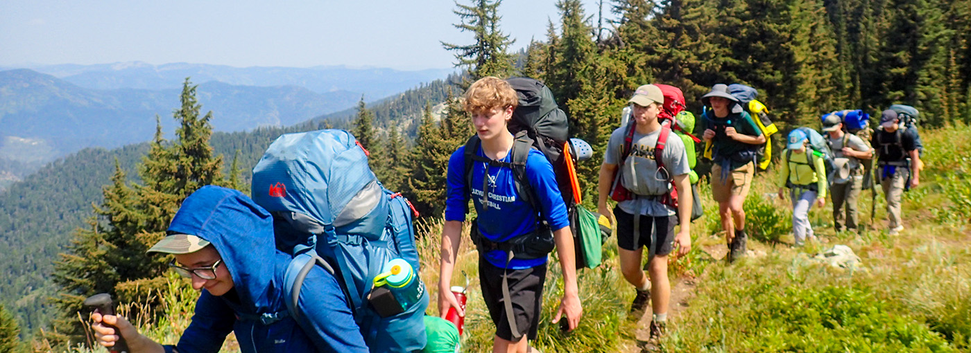 A line of hikers with large packs walk along a forested ridge of a mountain. 