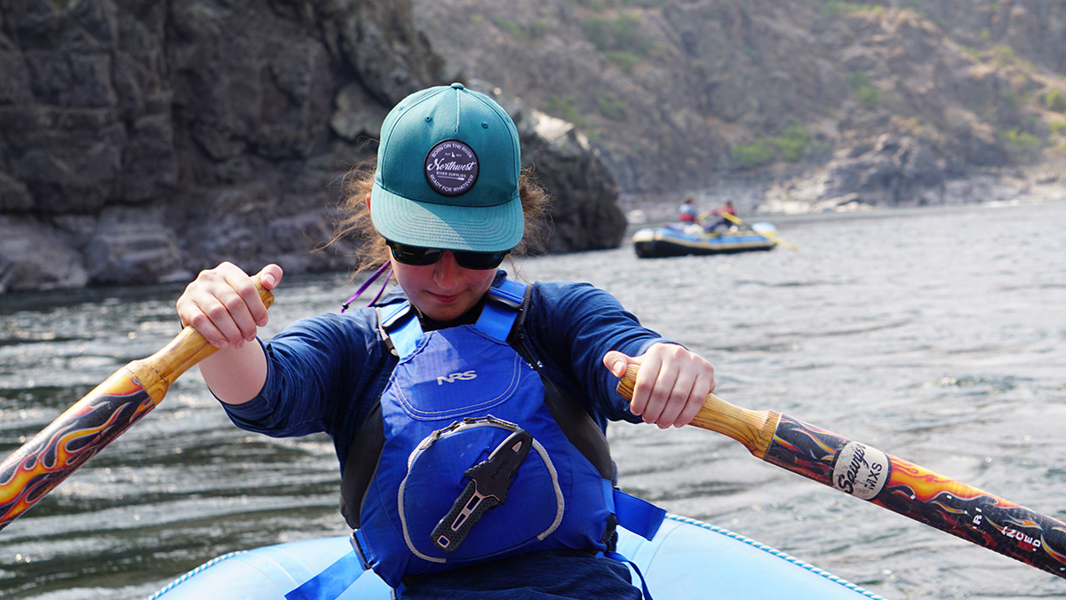 A woman in a blue hat and safety vest rows a raft through a canyon. 