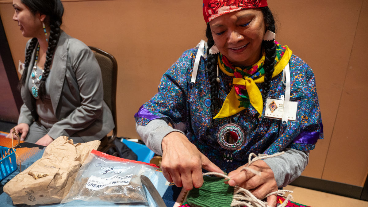 A woman in traditional Nez Perce clothing hand weaves a green digging bag.