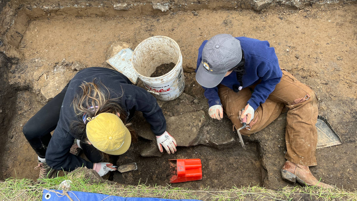 Two students use trowels as they dig dirt in a rectangular pit.